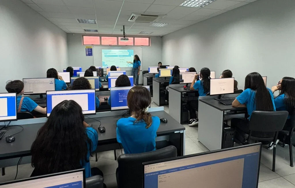 Classroom with students in blue shirts working on computers and a teacher presenting.